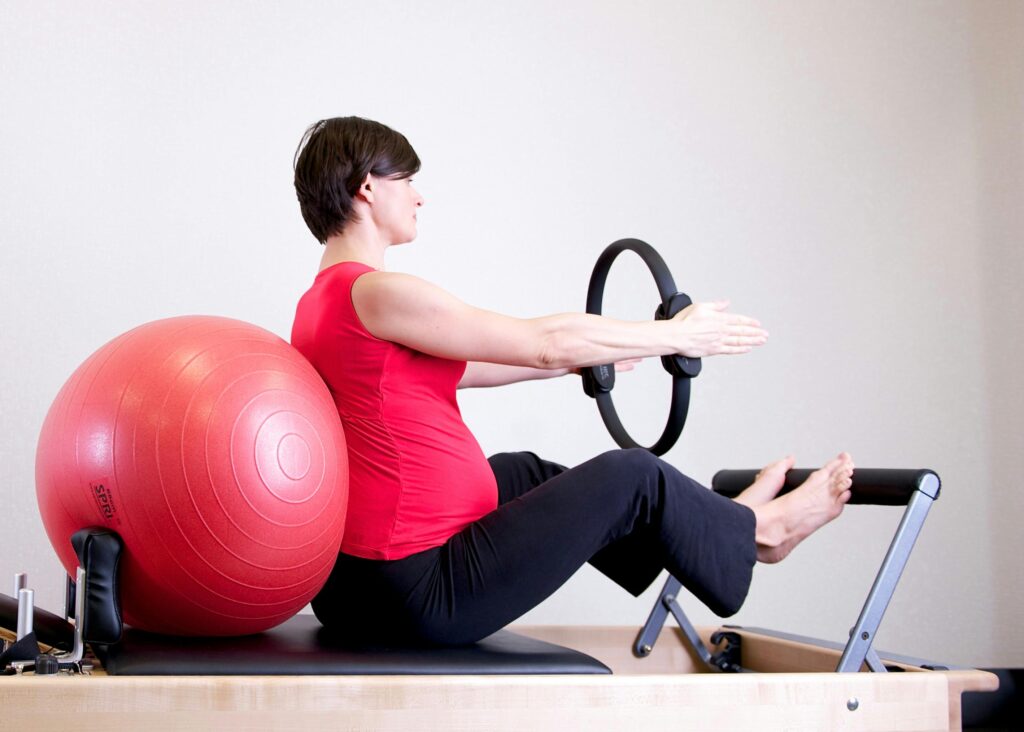 A pregnant woman in a red top practicing Pilates using a fitness ring and exercise ball in a gym setting.